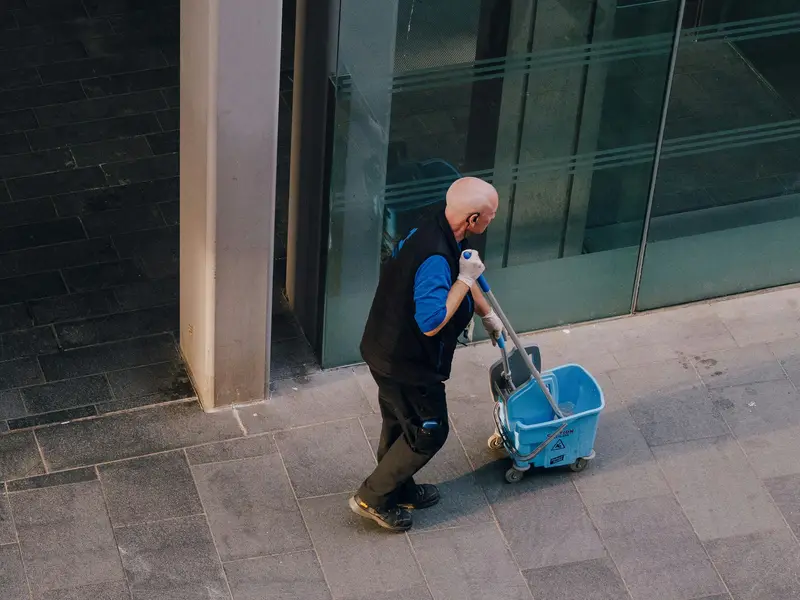 A janitor pulling a mop bucket and cleaning cart along the outside of a building