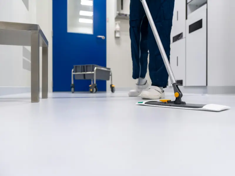 A cleaner in scrubs mopping the white floor of a clinical corridor