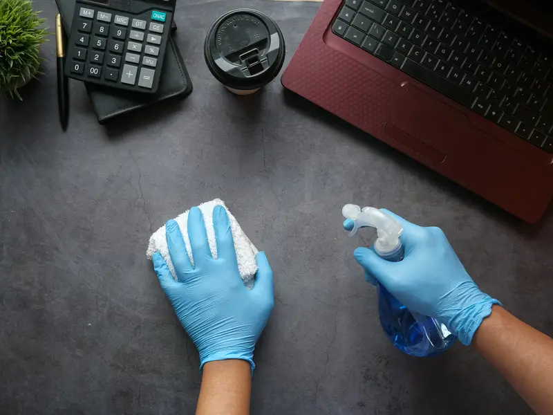 Gloved hands cleaning a desk around a keyboard and phone with a spray bottle and cloth